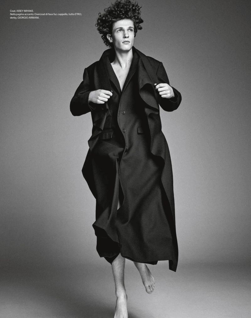 Monochrome portrait of a young male model with curly hair wearing a long dark coat against a plain grey backdrop.