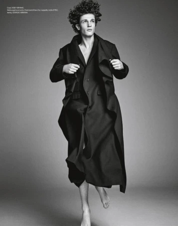 Monochrome portrait of a young male model with curly hair wearing a long dark coat against a plain grey backdrop.