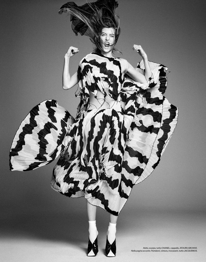 Model mid-air wearing a flowing patterned dress and white socks against a grey studio backdrop