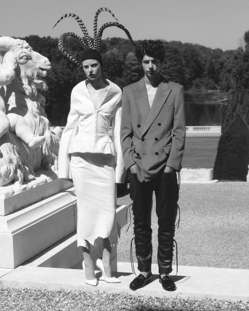 Two models posing outdoors near a stone sculpture and park landscape for V Magazine editorial