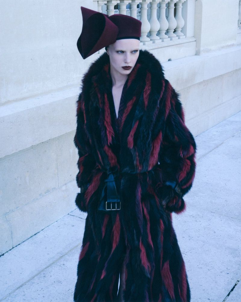 Model wearing a black and red fur coat and sculptural hat standing against a classical stone building exterior.