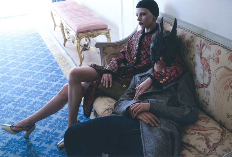 Two models seated on an ornate patterned sofa in an interior, one wearing a patterned coat and black cap, the other in a grey coat and mask.