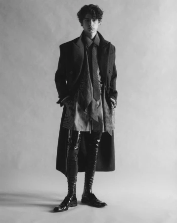 Monochrome fashion editorial portrait of a male model wearing a tailored coat and glossy thigh-high boots against a grey studio backdrop.