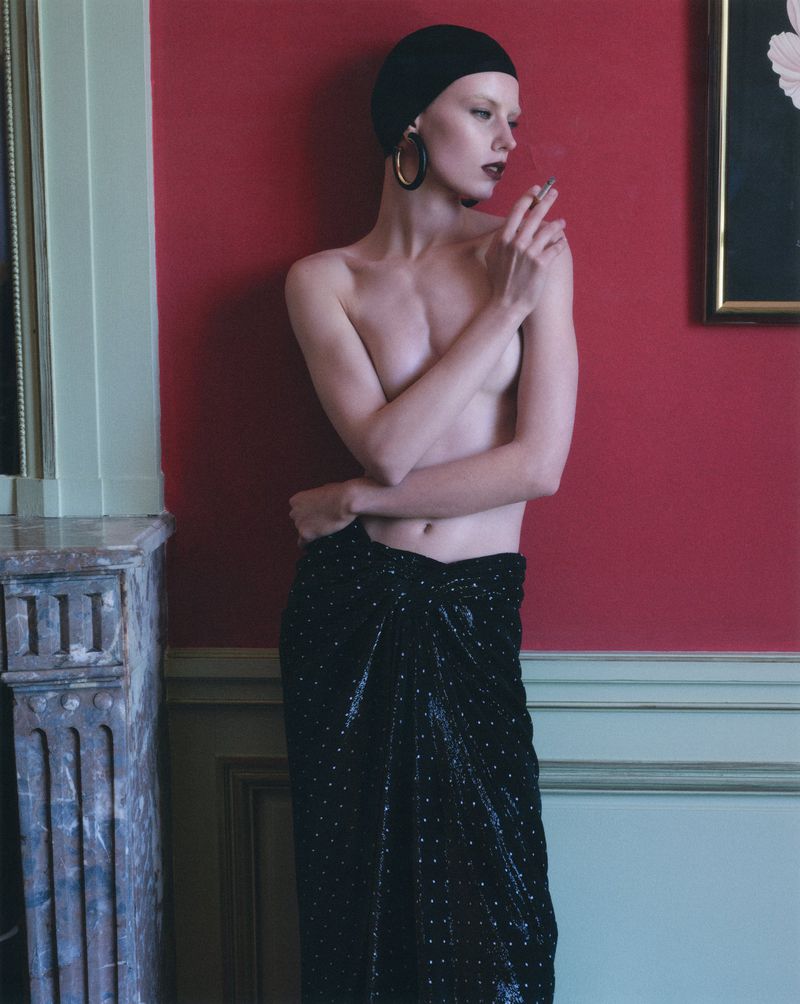 Model in black swim cap and skirt holding a cigarette against a red wall for V Magazine