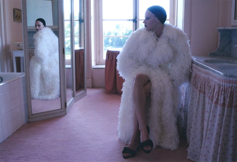 Model wearing white furry coat and dark cap seated in a pink-carpeted room with a tall mirror reflecting her figure.