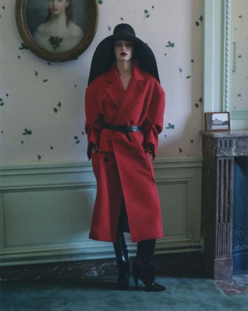 Model wearing a red coat and wide black hat standing against a patterned wall for V Magazine