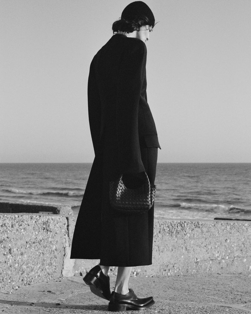 Model wearing a black coat and hat holding a woven handbag against an ocean background