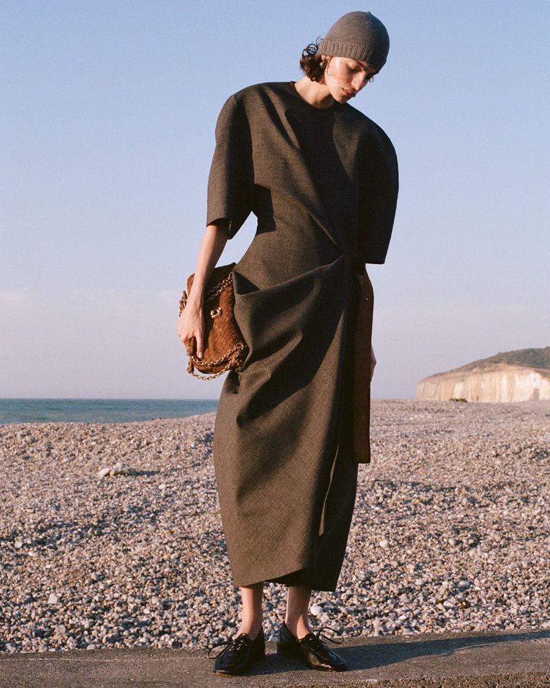 Model wearing a structured dark dress and beanie holding a brown textured handbag on a pebbled beach.