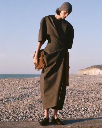 Model wearing a structured dark dress and beanie holding a brown textured handbag on a pebbled beach.