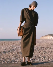 Model wearing a structured dark dress and beanie holding a brown textured handbag on a pebbled beach.