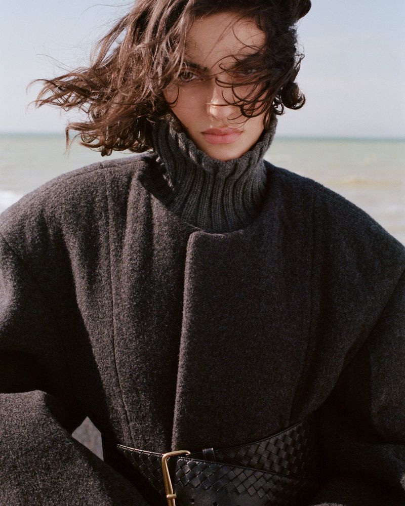 Model with dark hair blowing in the wind wearing a heavy grey wool coat and ribbed turtleneck against a coastal horizon background.