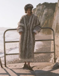 Model standing outdoors near a railing against a rocky coastal cliff and ocean shoreline wearing a fuzzy textured coat and matching hat