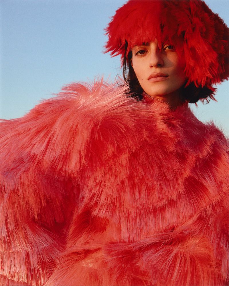 Model wearing a vibrant red fringed hat and textured coat against a clear blue sky