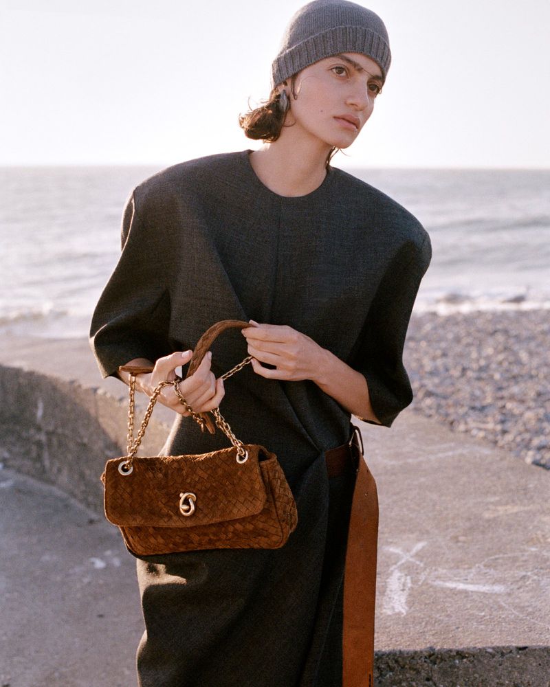 Model wearing a dark grey dress and beanie holding a brown suede chain handbag outdoors by the coast for Elle Canada.