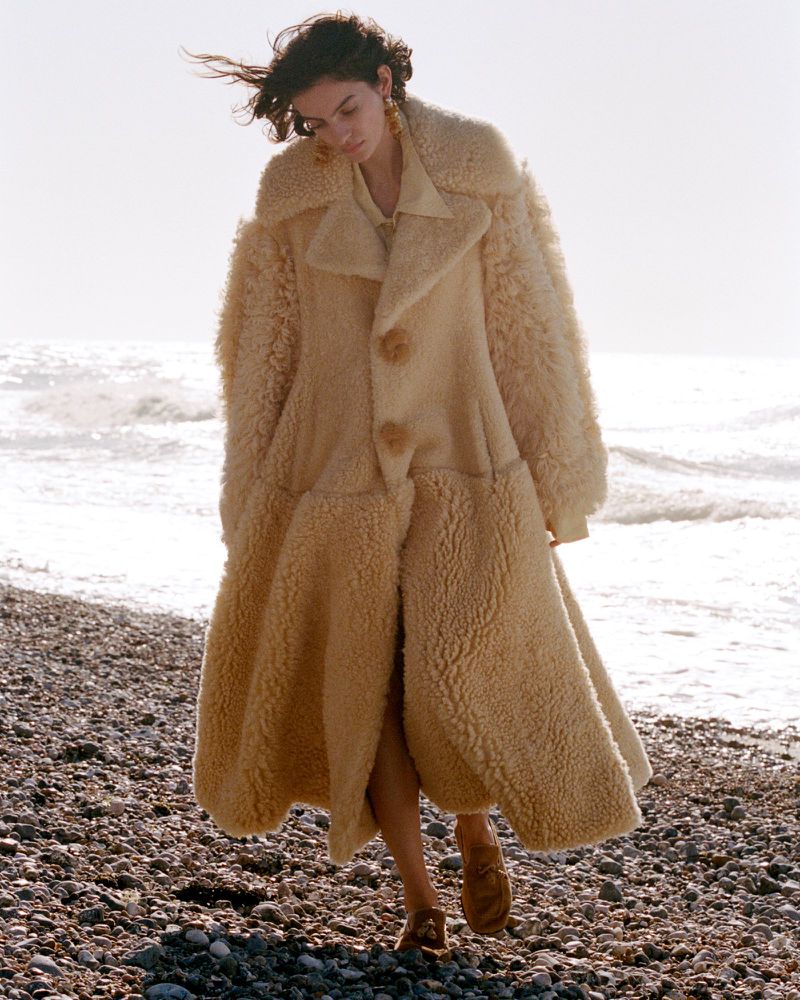 Model in a thick cream shearling coat walking along a pebble beach with ocean waves behind.