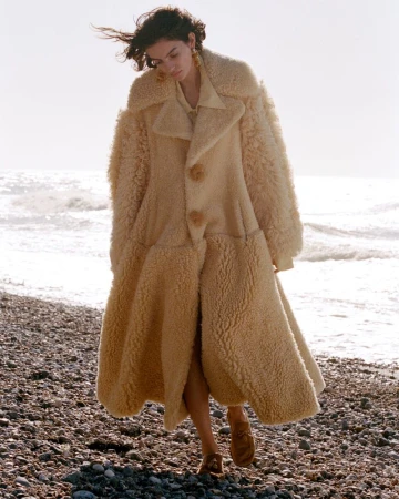 Model in a thick cream shearling coat walking along a pebble beach with ocean waves behind.