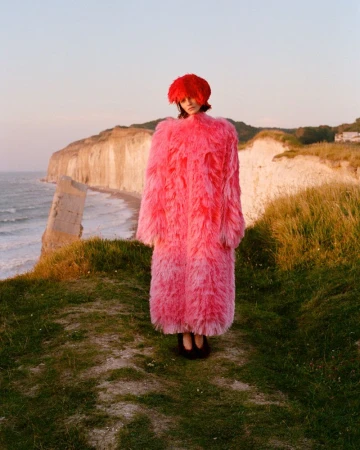 Model standing on coastal cliffs wearing a vibrant pink fringed coat and matching red headwear for Elle Canada editorial.