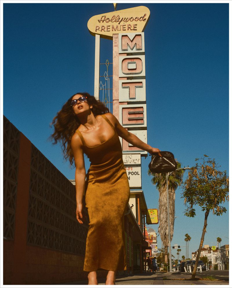Adria Arjona wearing a brown dress and sunglasses holding a black handbag under a motel sign