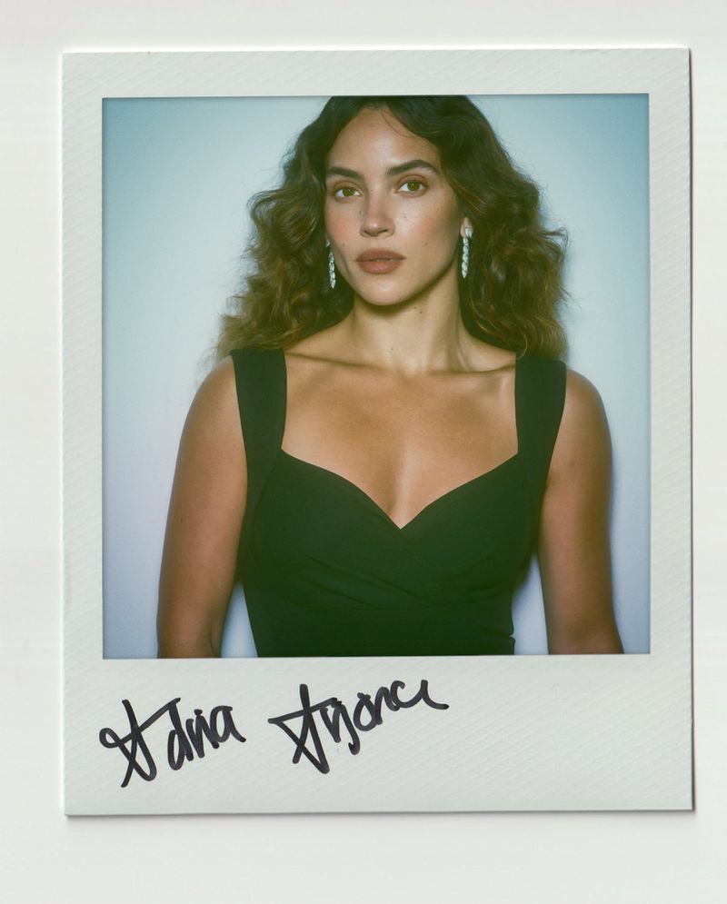 Adria Arjona in a black dress with long wavy hair against a white background within a Polaroid frame featuring handwritten signature