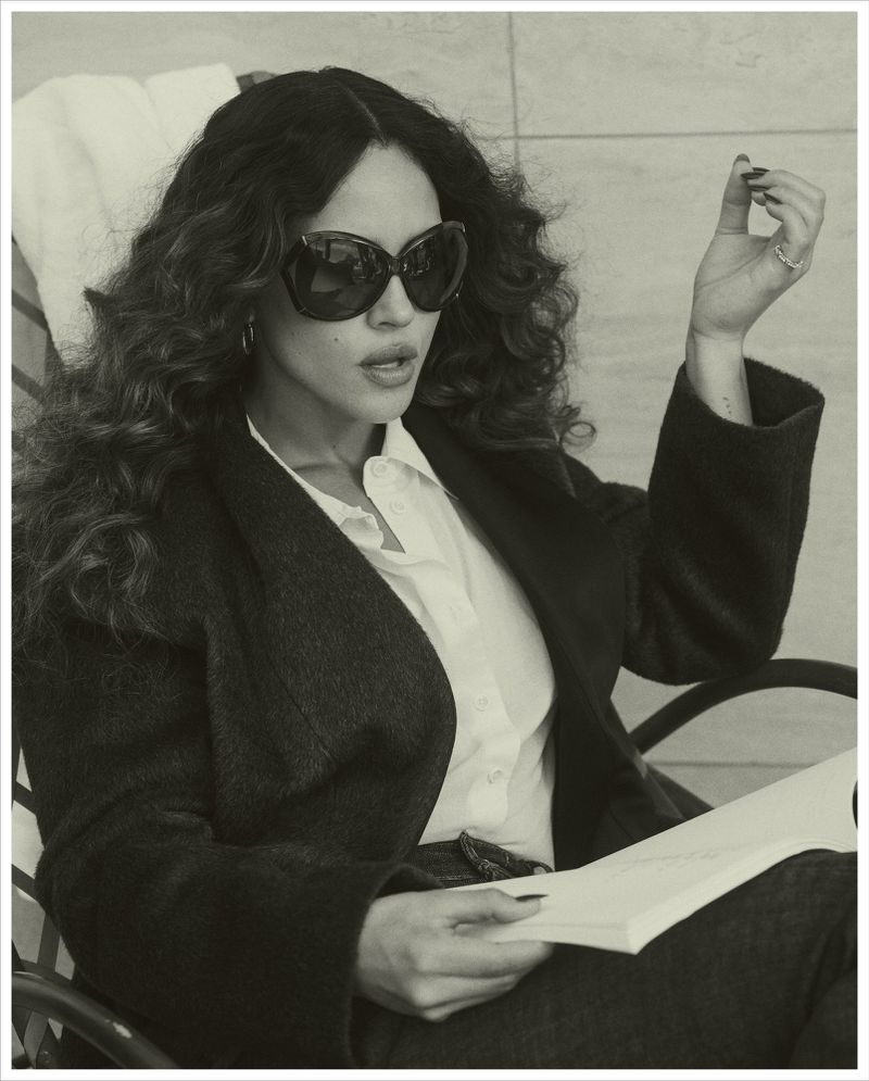 Adria Arjona wearing oversized sunglasses and a dark coat while seated outdoors for Vogue Mexico
