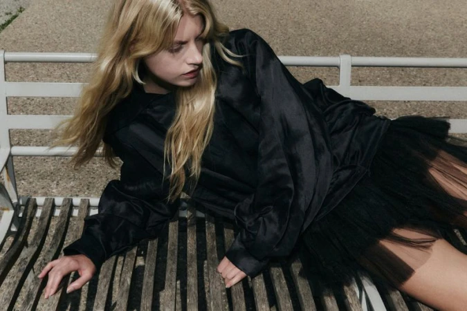 Blonde woman wearing a black satin blouse and tulle skirt reclining on a wooden park bench.