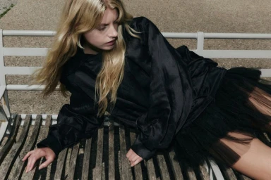 Blonde woman wearing a black satin blouse and tulle skirt reclining on a wooden park bench.