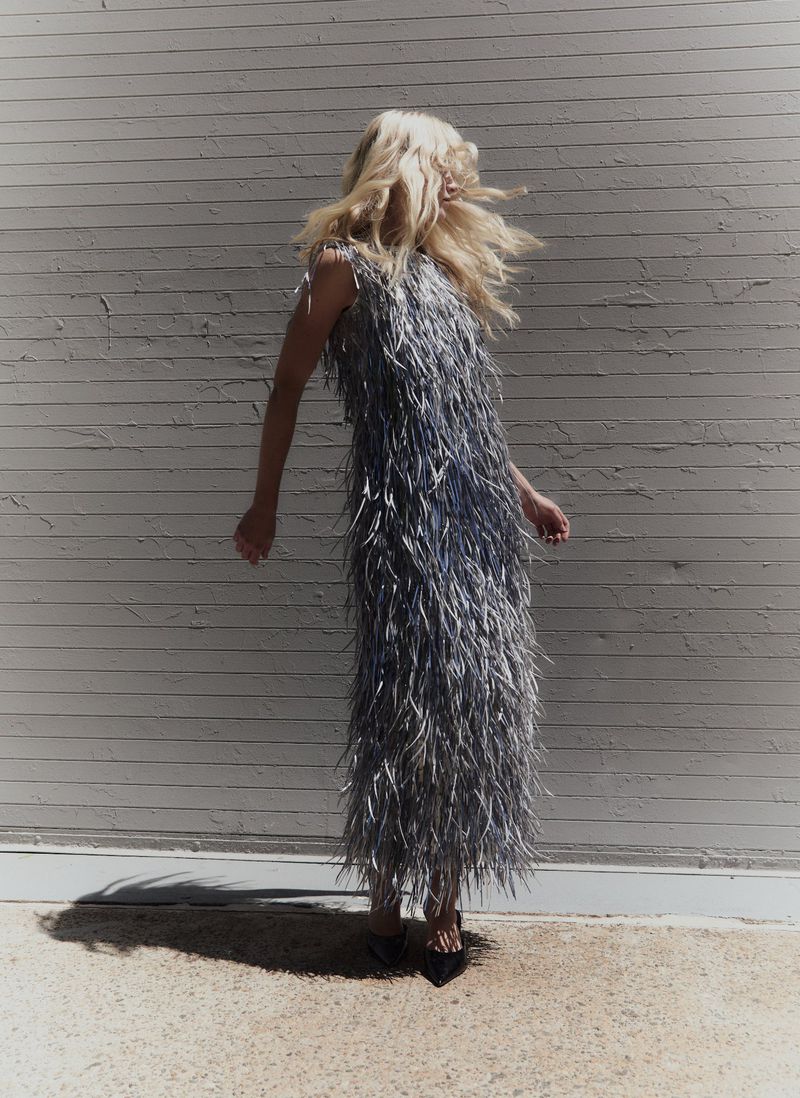 Blonde model in a heavily textured grey fringed dress standing against a horizontal slat wall in harsh sunlight.