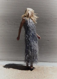 Blonde model in a heavily textured grey fringed dress standing against a horizontal slat wall in harsh sunlight.