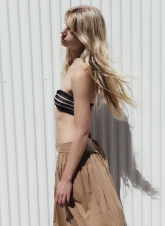 Model with long blonde hair wearing a black tube top and tan skirt against a white corrugated panel background