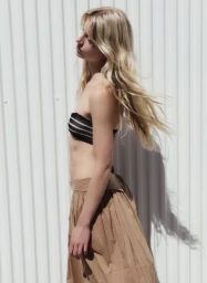 Model with long blonde hair wearing a black tube top and tan skirt against a white corrugated panel background