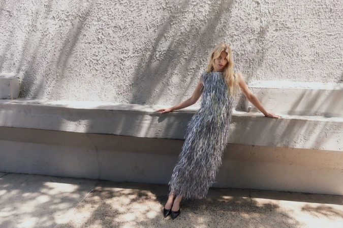 Model wearing a grey fringed dress leaning against a textured concrete wall with tree shadow patterns