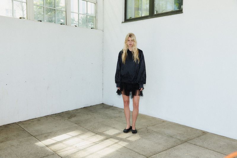 Model wearing dark long-sleeve top and sheer black skirt standing inside a white room with concrete floor