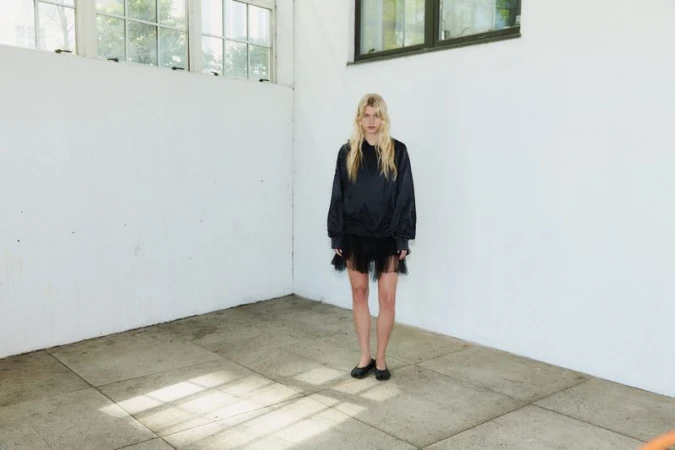 Model wearing dark long-sleeve top and sheer black skirt standing inside a white room with concrete floor