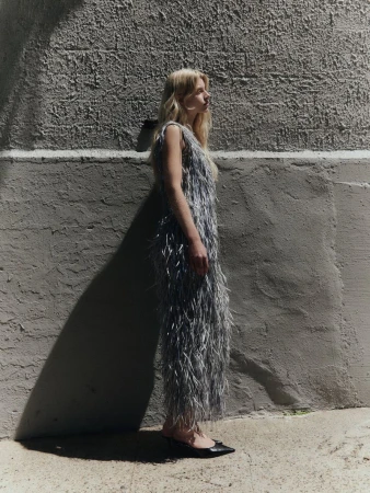Model wearing a textured fringed dress posing against a rough concrete wall in natural light.