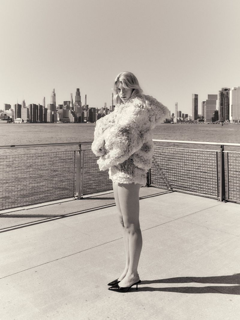 Model wearing a textured fluffy jacket standing on a waterfront pier with a city skyline in the background.