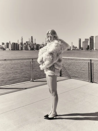 Model wearing a textured fluffy jacket standing on a waterfront pier with a city skyline in the background.