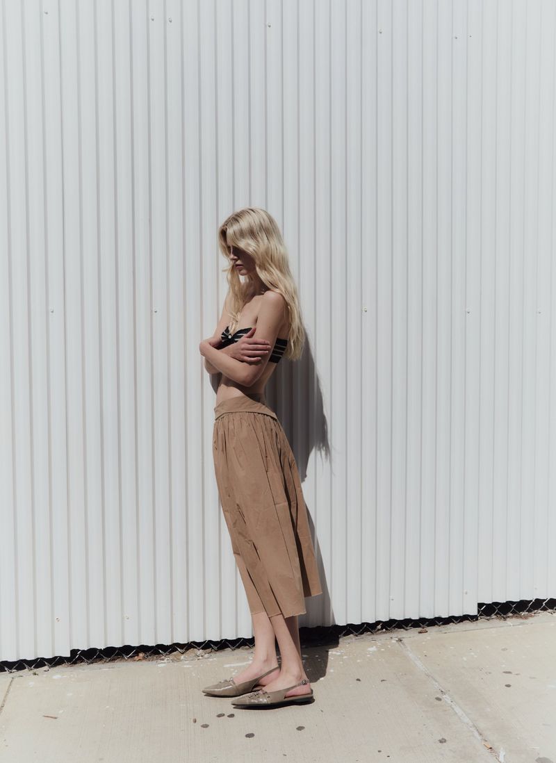 Model standing in profile against a white corrugated metal wall wearing a tan skirt and flat shoes