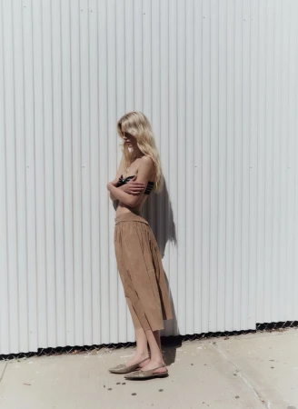 Model standing in profile against a white corrugated metal wall wearing a tan skirt and flat shoes