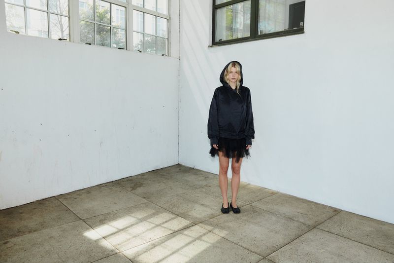 Model wearing a black hooded jacket and dark skirt standing in an empty room with white walls and a grid-paned window.