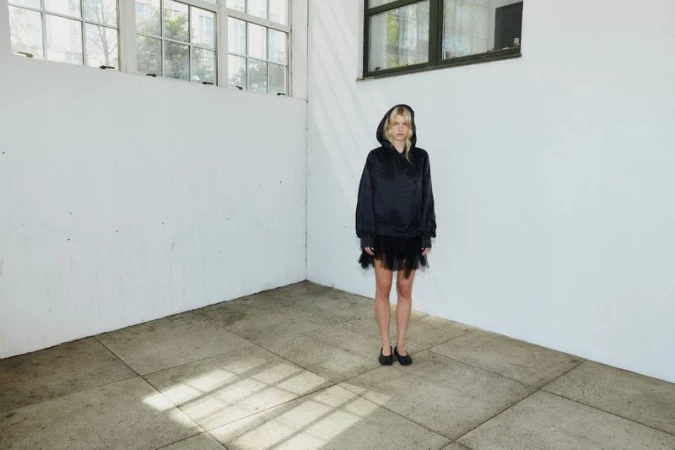 Model wearing a black hooded jacket and dark skirt standing in an empty room with white walls and a grid-paned window.