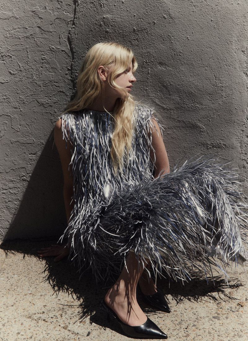 Blonde model wearing a textured silver fringed dress seated against a rough concrete wall outdoors