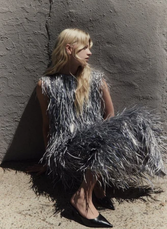 Blonde model wearing a textured silver fringed dress seated against a rough concrete wall outdoors