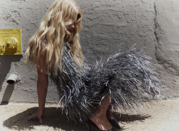 Blonde model in a textured grey feathered dress seated against a textured concrete wall with a yellow fire hydrant.