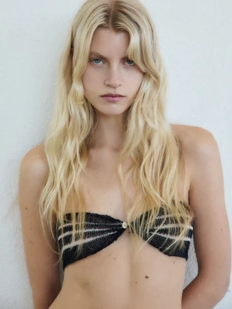 Close-up portrait of a blonde woman wearing a black strapless knit top against a plain textured wall.