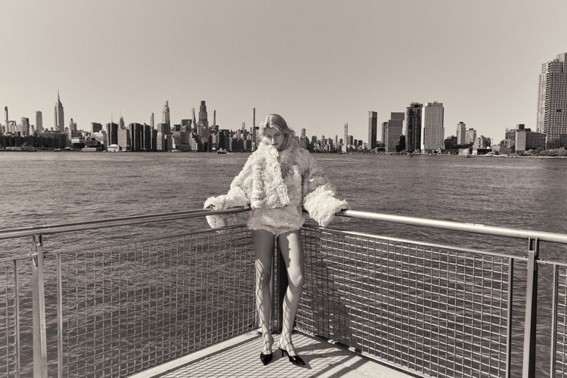 Model wearing textured white top leaning on a metal railing with a city skyline and water view in the background