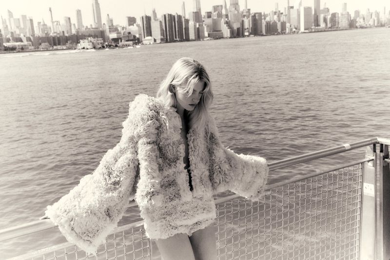 Blonde female model leaning on a metal railing over water with a distant city skyline in the background for Quiet Motion
