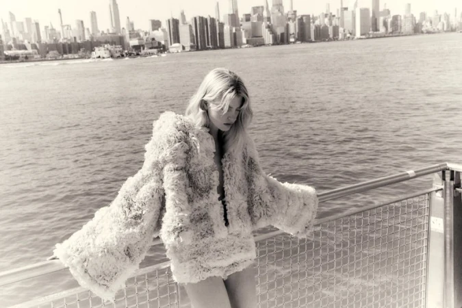 Blonde female model leaning on a metal railing over water with a distant city skyline in the background for Quiet Motion