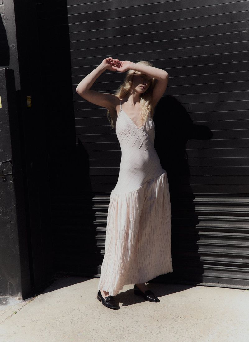Blonde female model wearing a cream slip dress and black loafers standing against a dark corrugated metal door.