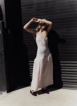 Blonde female model wearing a cream slip dress and black loafers standing against a dark corrugated metal door.