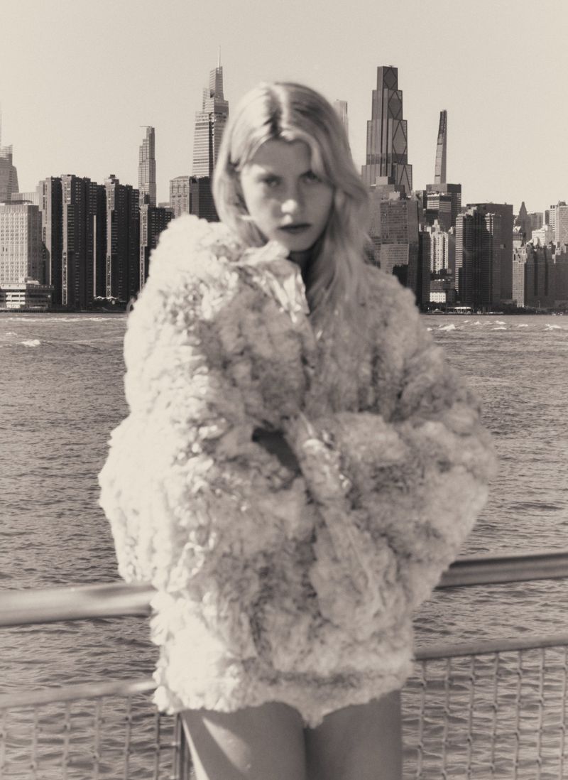 Model wearing a textured white coat standing against a city skyline and waterfront view
